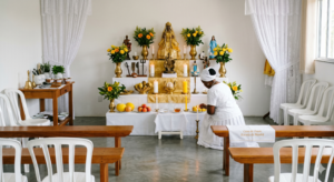 Interior iluminado e sereno do terreiro Casa da Oxum Estrela da Manhã em Guarulhos. O altar está impecavelmente limpo e organizado, decorado com tecidos brancos e dourados, flores amarelas, velas acesas e imagens sagradas. Uma mulher negra, vestida com as roupas brancas tradicionais da Umbanda, está ajoelhada cuidando do altar com profundo zelo e respeito.